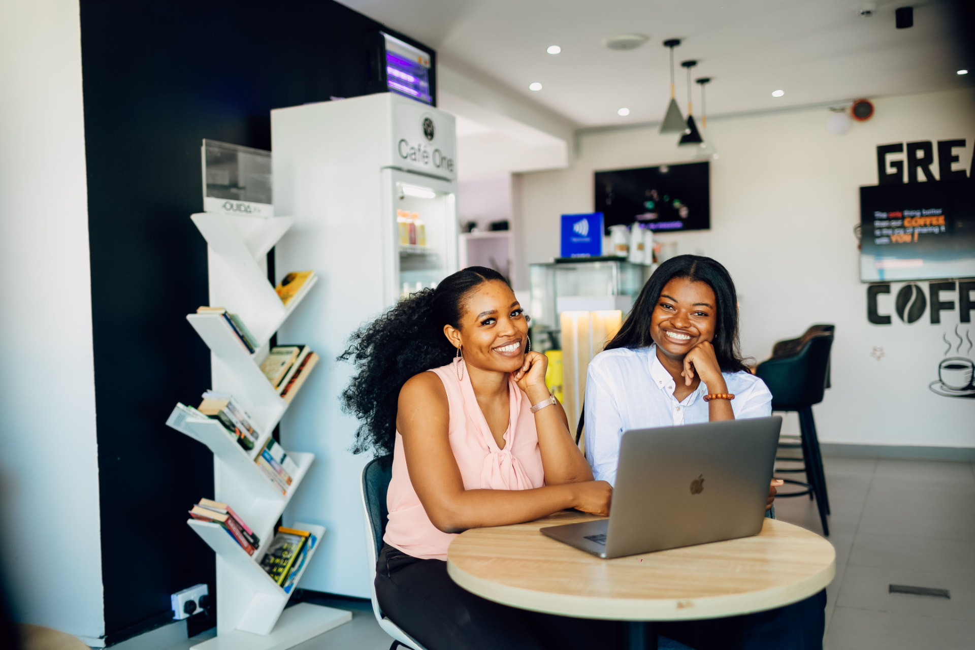 Two women sitting together at a small table with a laptop in a bright café space